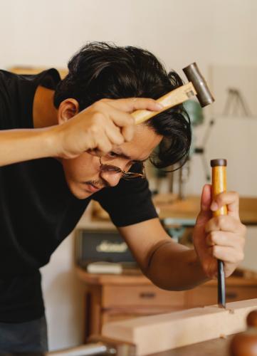 Young male artisan shaping wooden plank with hammer and chisel.