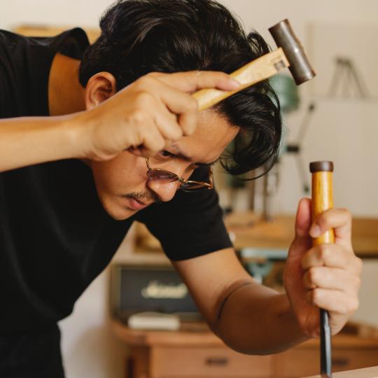 Young male artisan shaping wooden plank with hammer and chisel.