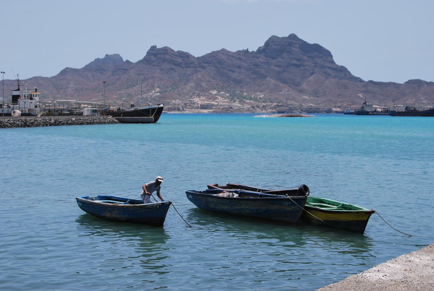 A fisher tying a small boat in the harbour, with Mindelo and its mountains across the water