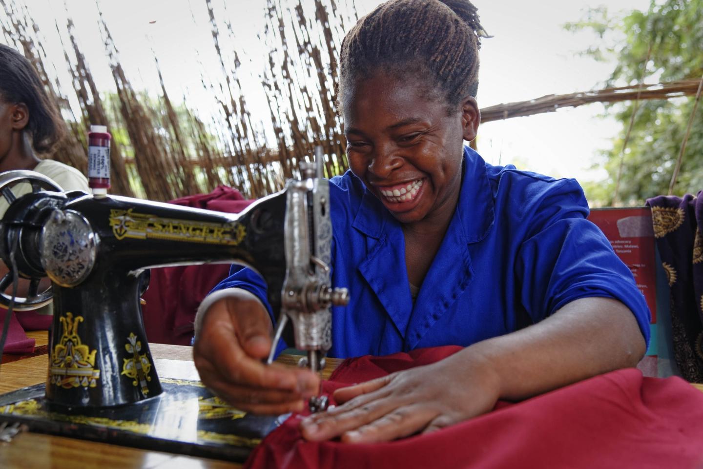 A smiling seamstress in a blue work coat sewing red fabric at a manual sewing machine