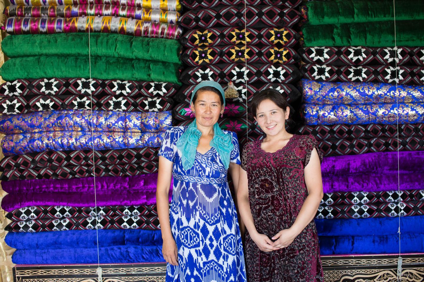 Two women standing in front of stacked colourful handwoven textiles